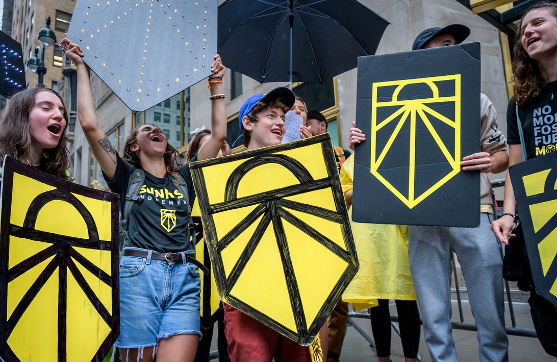 Klimatdemonstration med unga aktivister som håller gulsvarta sköldformade skyltar med en solsymbol. Ungdomarna marscherar i en stadsmiljö med allvarliga ansiktsuttryck som visar allvaret i deras klimataktivism.